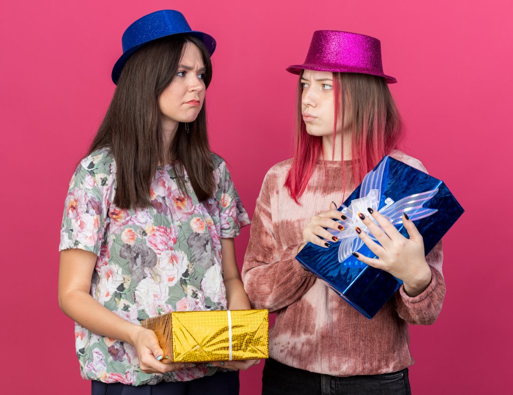 unpleased-girls-wearing-party-hat-holding-gift-boxes-look-at-each-other-isolated-on-pink-wall.jpg