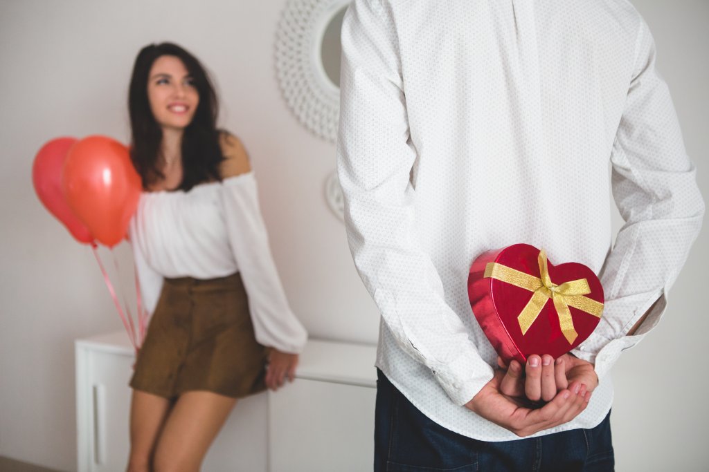 girl-holding-balloons-with-heart-shape-while-her-boyfriend-has-a-gift-for-her-to-the-back.jpg